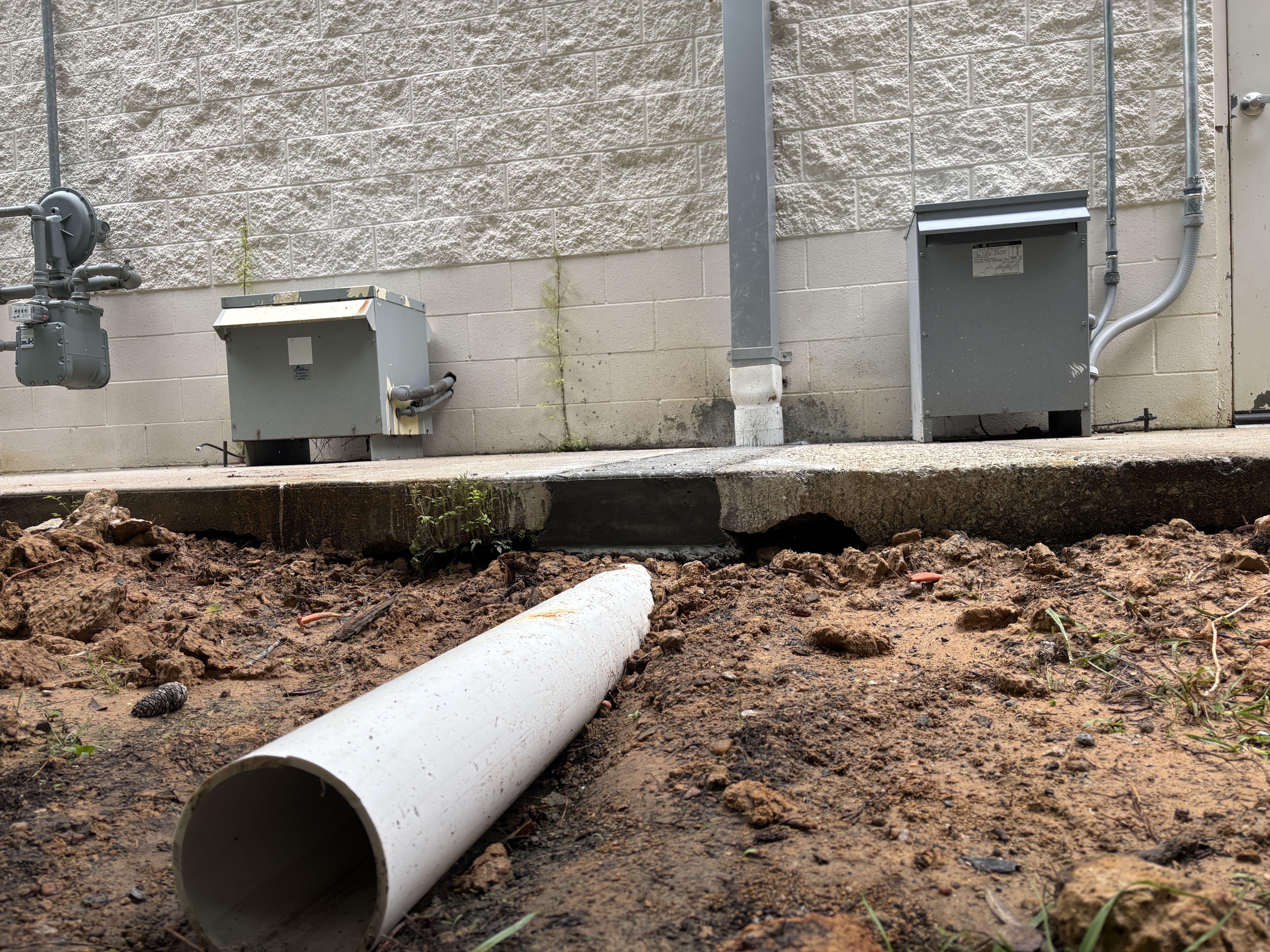 Open trench alongside a commercial building with PVC storm drain pipe staged, showing soil washed out from under the concrete walkway